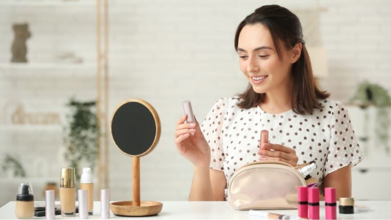 Beautiful young woman with cosmetic bag and lipsticks doing makeup at table in dressing room