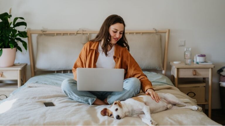 Young student e-learning on laptop at home, cute small dog besides. Businesswoman working remotely, sitting on bed. Homeoffice for single woman living alone.