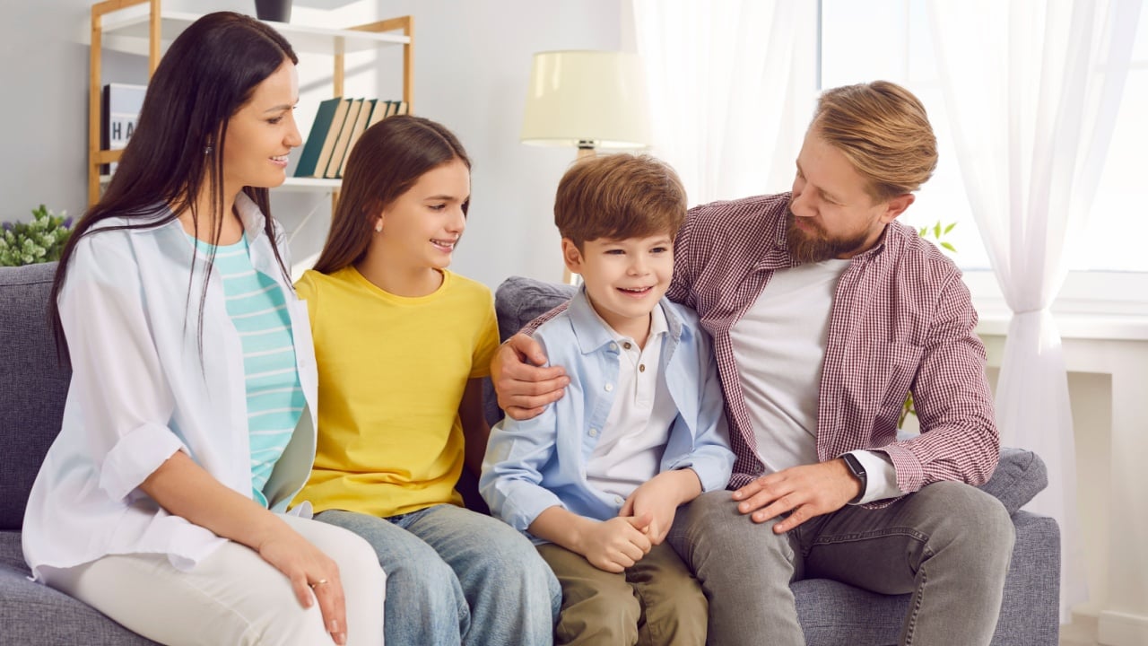 Smiling young happy family of four with two kids boy and girl sitting on the sofa at home spending weekend together. Portrait of cheerful parents with children. Family time and leisure concept.