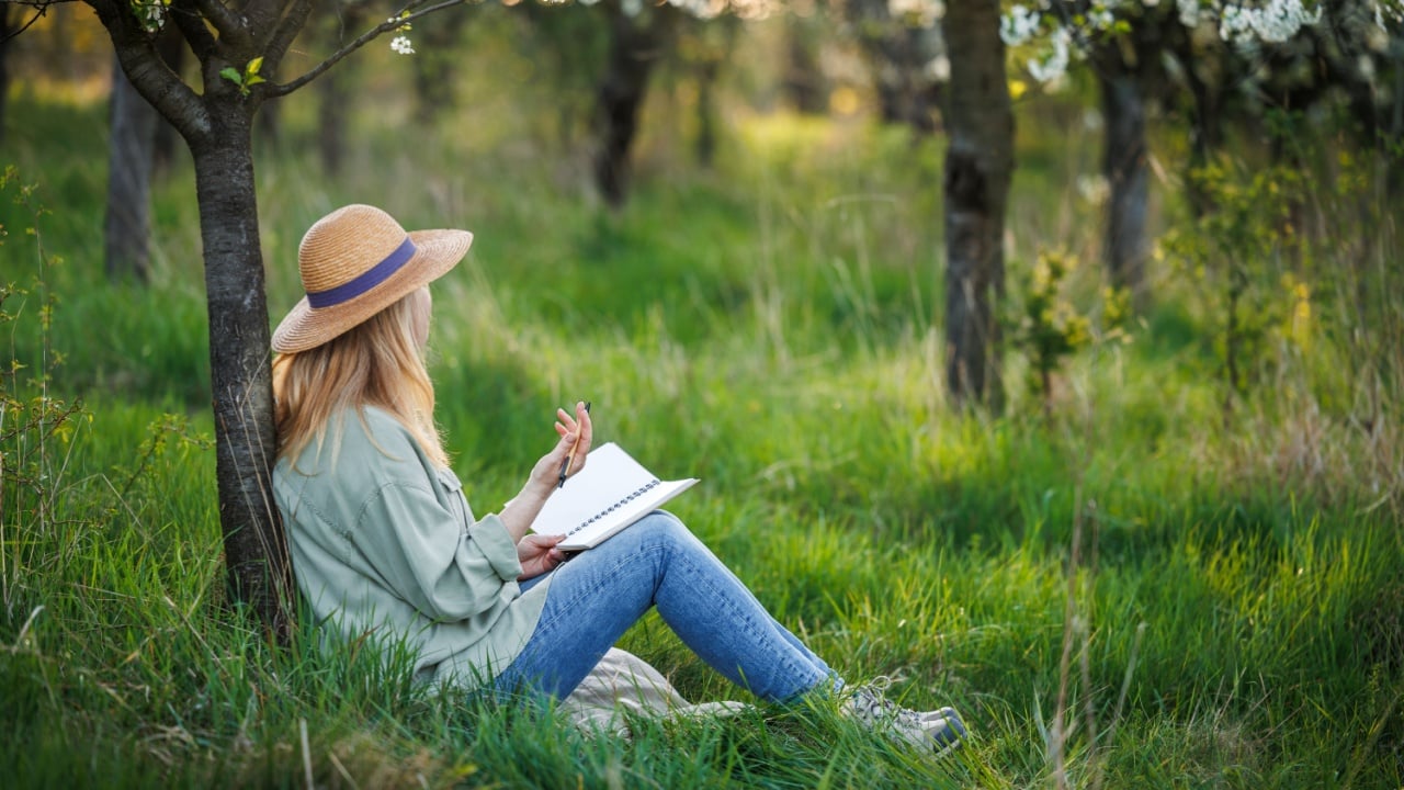Woman is writing diary and resting in blossoming orchard. Mindfulness and reconnect the moment in spring nature