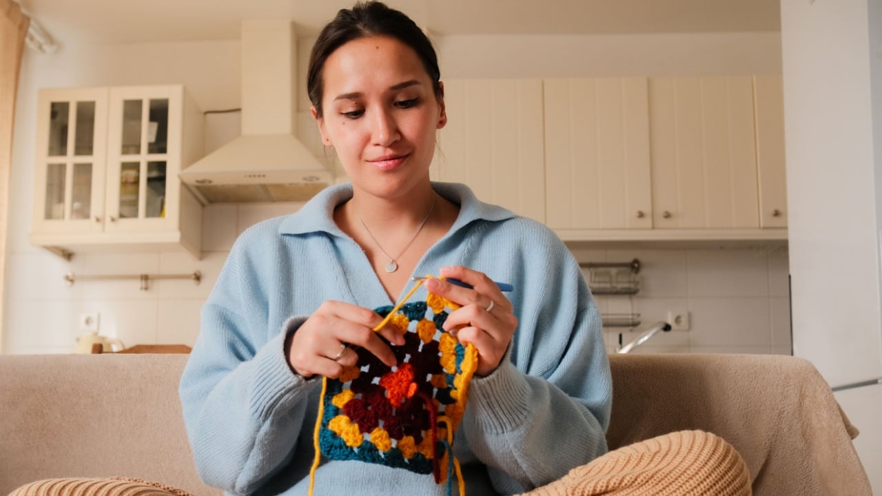 Young woman knitting crocheting with colored yarn granny square at home. Woman doing needlework, home hobbies.
