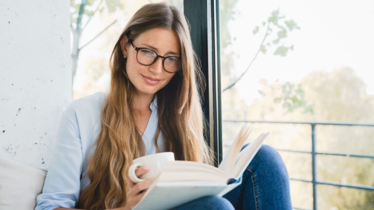 Smart Caucasian woman reading a novel at home by the window. Freelancer student female reading a book, preparing for exam project, doing homework, inspired with the plot