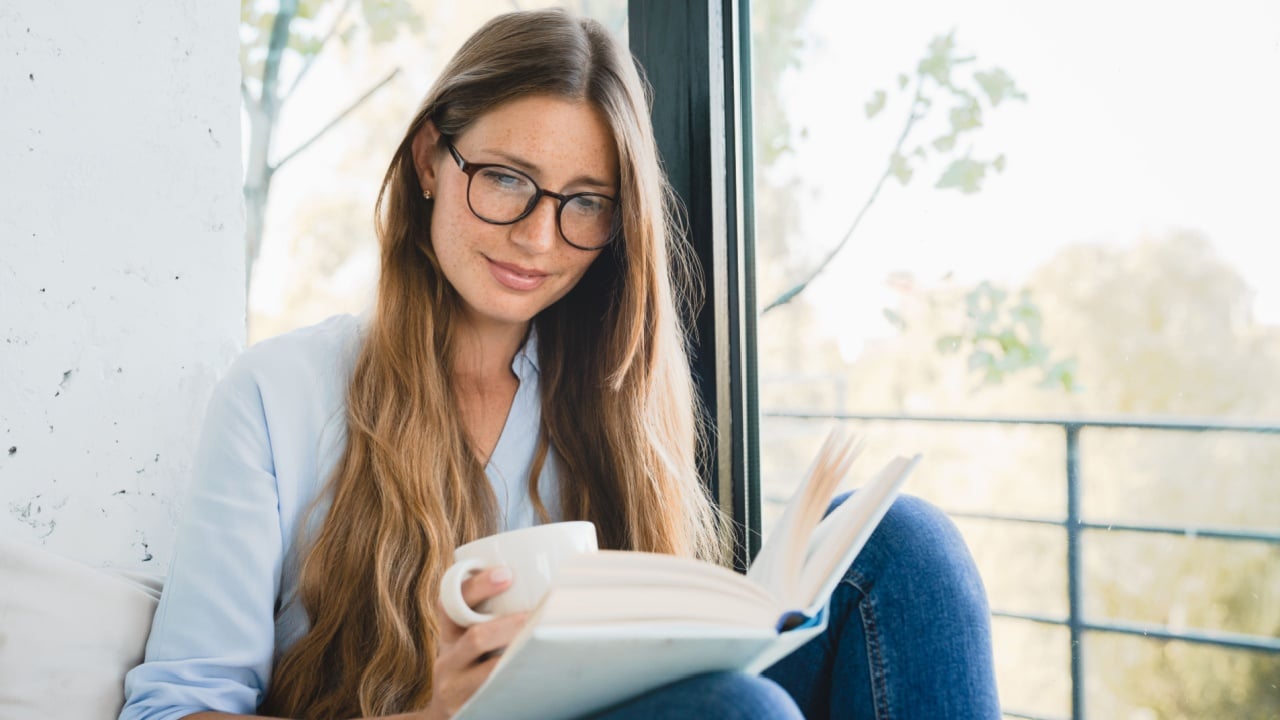 Smart Caucasian woman reading a novel at home by the window. Freelancer student female reading a book, preparing for exam project, doing homework, inspired with the plot