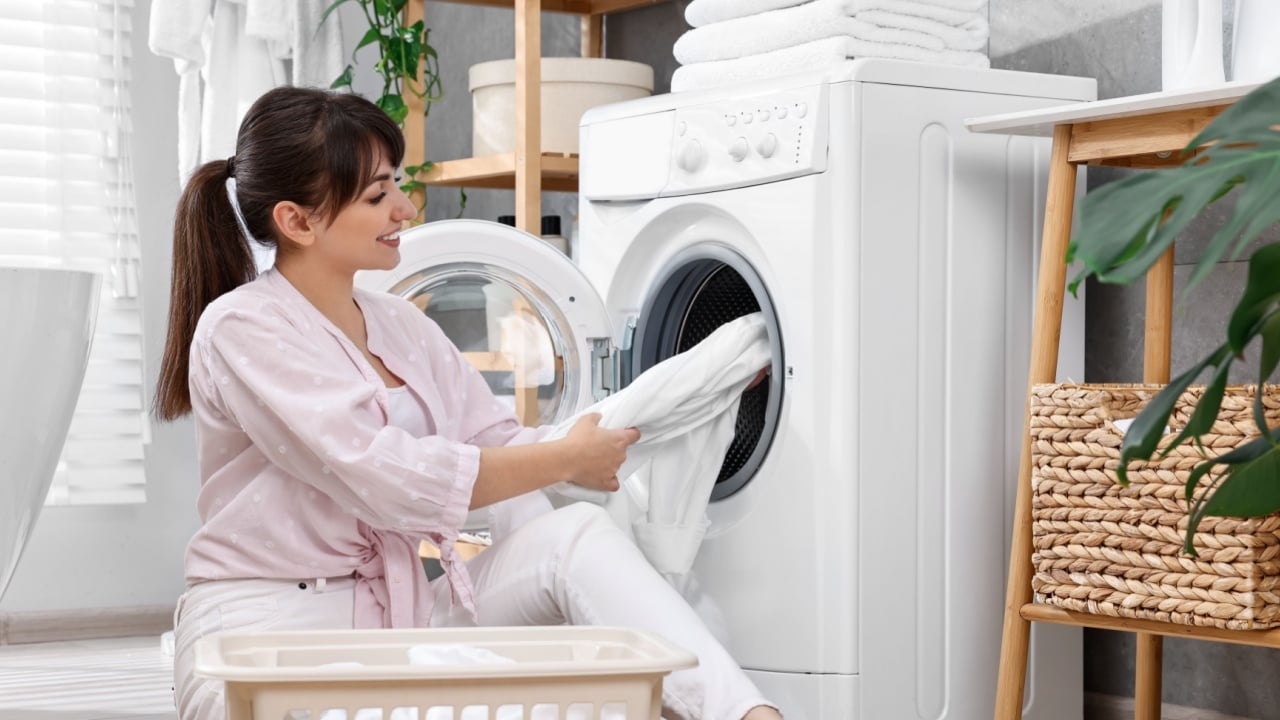 Happy young housewife putting laundry into washing machine at home