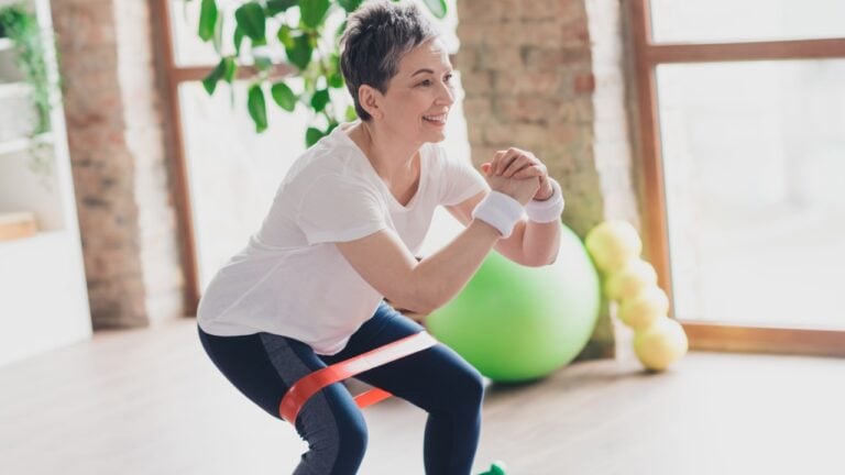 Photo of attractive senior woman resistance band squats sportswear home workout spacious bright living room sport concept