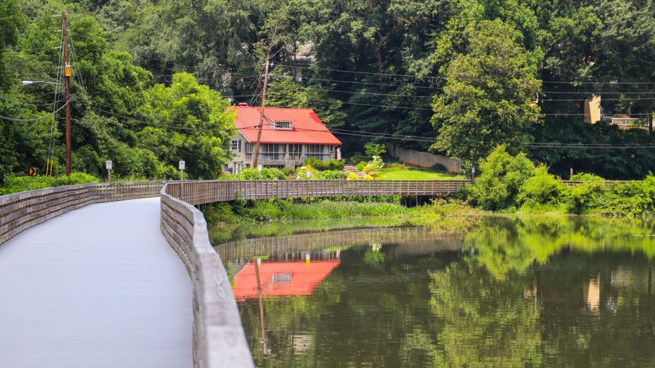 a house with a red roof along the Chattahoochee river with a long winding boardwalk with a wooden fence with lush green trees and plants at Roswell Riverwalk Boardwalk in Roswell Georgia