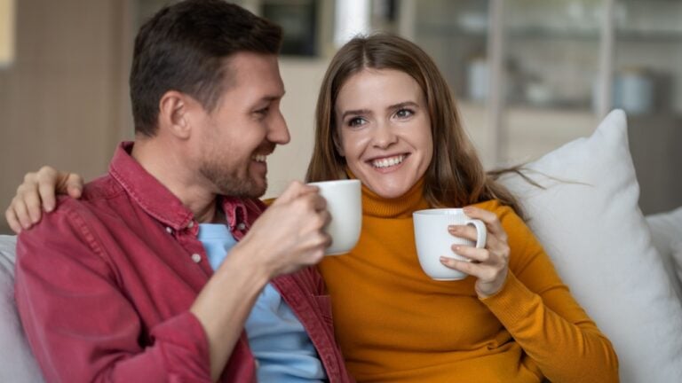 Loving couple man woman drink tea or coffee on cozy sofa at home, enjoy talking spending lazy weekend. Affectionate spouses wife husband bonding have comfort pastime. Harmonious marriage relationship.