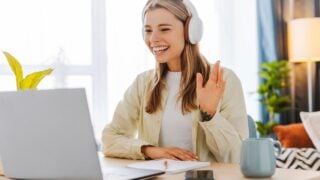 Young woman is using laptop for video communication while sitting at table and working from home waving hand. Online communication