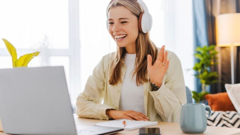Young woman is using laptop for video communication while sitting at table and working from home waving hand. Online communication