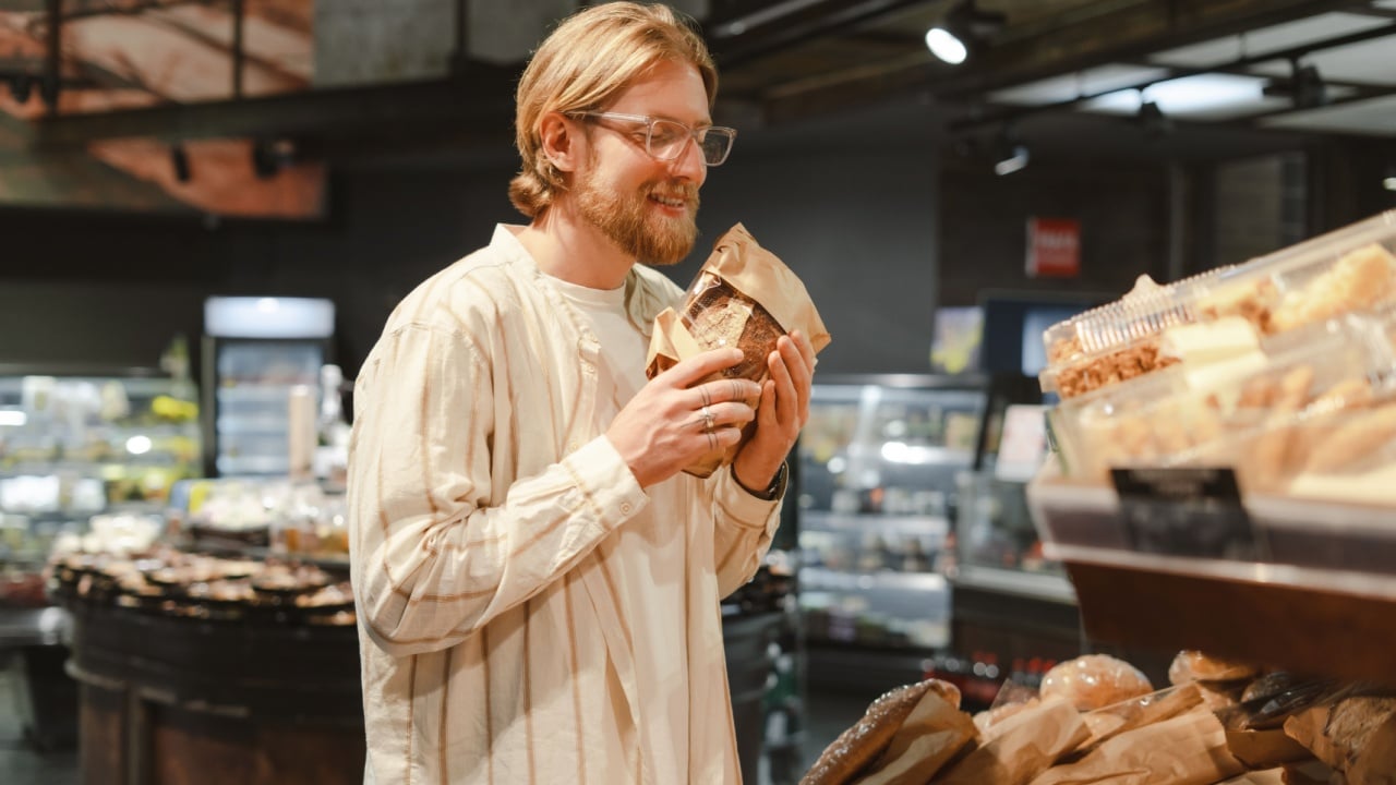 Man with glasses choosing bread in a bakery section of a grocery store, surrounded by various types of loaves. The aroma is tempting, making him hungry