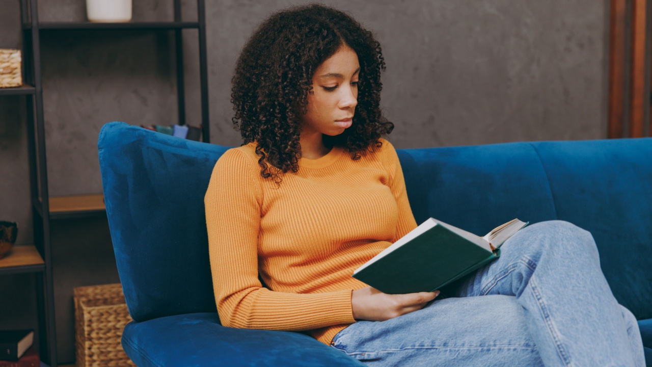 Side view little smart kid teen girl wear casual clothes reading novel paper book sits on blue sofa couch stay at home hotel flat rest relax spend free spare time in living room indoor.
