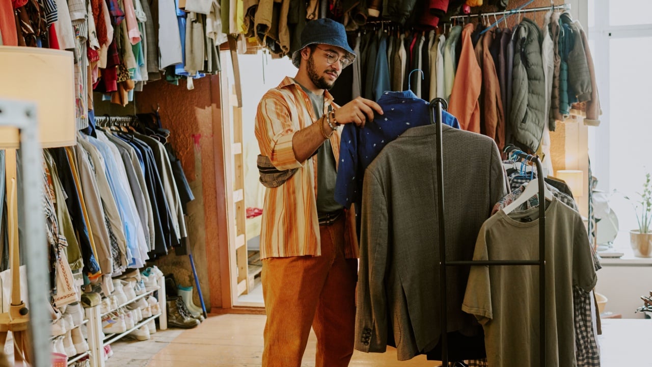 Person browsing clothes in a cozy vintage thrift shop filled with various garments and accessories hanging on racks
