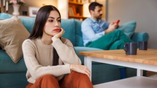 Young couple unhappy and angry after arguing at home, woman sitting sad and pensive while man using smart phone on the other side of the room