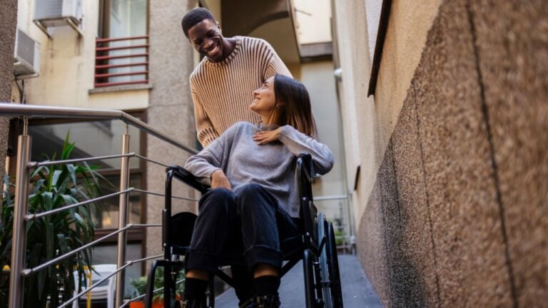 Smiling young man assists a woman in a wheelchair on a ramp outside a building. Young man helping woman in wheelchair outdoors with a smile