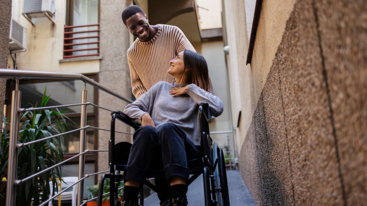 Smiling young man assists a woman in a wheelchair on a ramp outside a building. Young man helping woman in wheelchair outdoors with a smile