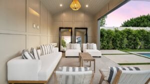 Modern outdoor living space with white furniture, wooden accents, and a view of lush greenery and a pool at sunset.