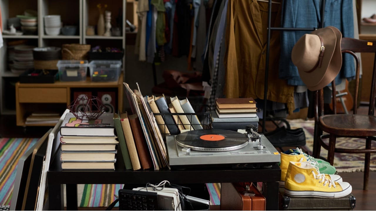 Several stacks of books, stereo record player on table standing by suitcases with sports shoes on top against racks with attire