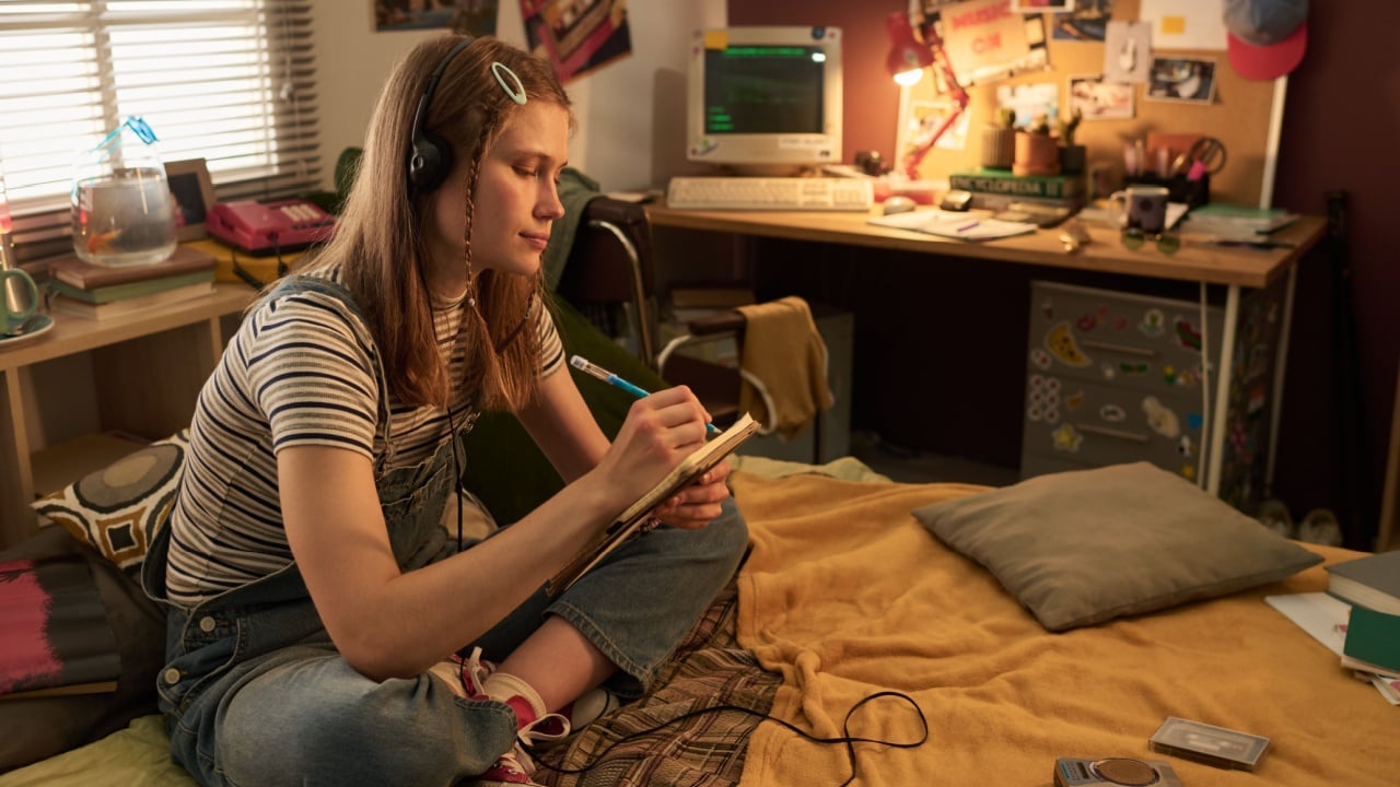 Young woman sitting on bed, writing in notebook while listening to music. Room filled with personal retro items and computer desk in background, creating nostalgic feel