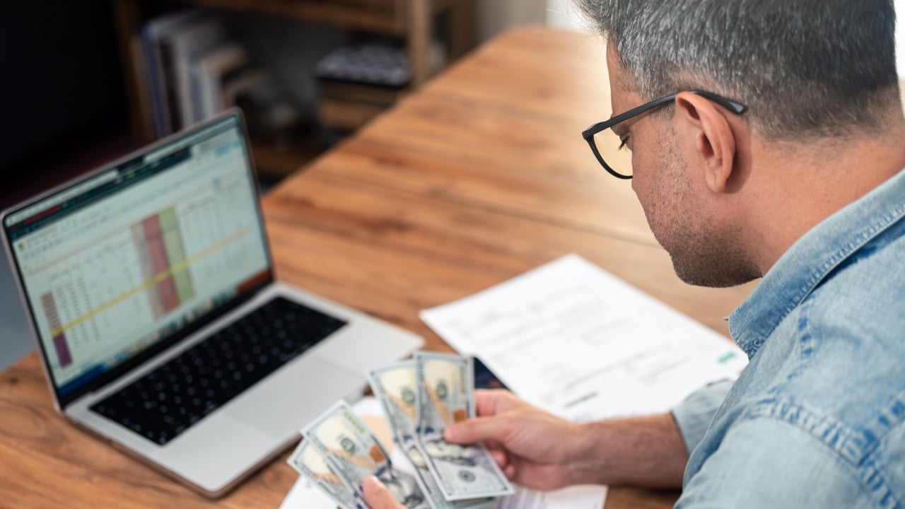 Doubted grey haired man looks at dollar banknotes over bills stack in commercial office, mature man thinks about money lack for household expenses at workplace