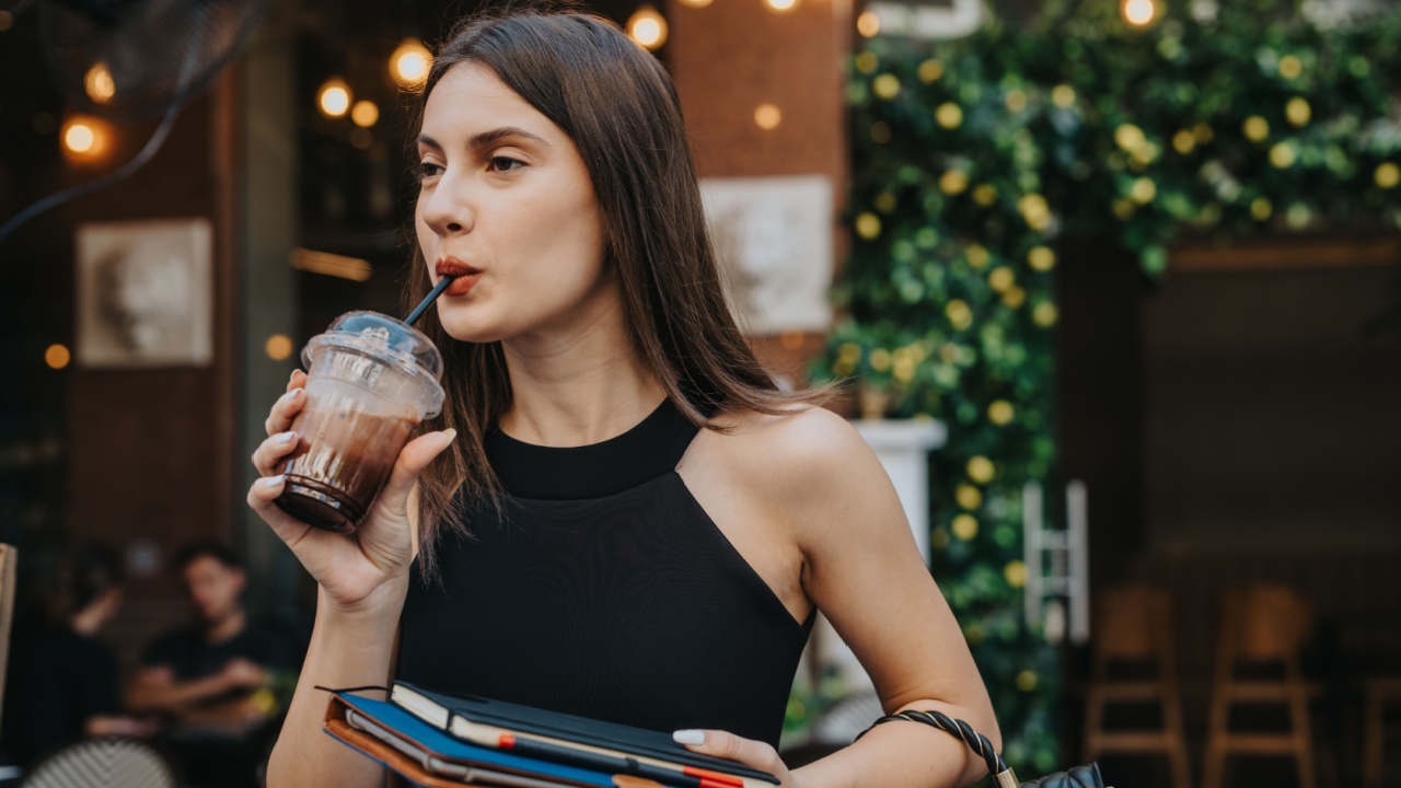 Fashionable young woman sipping iced coffee outside a stylish cafe while carrying notebooks and a black handbag.