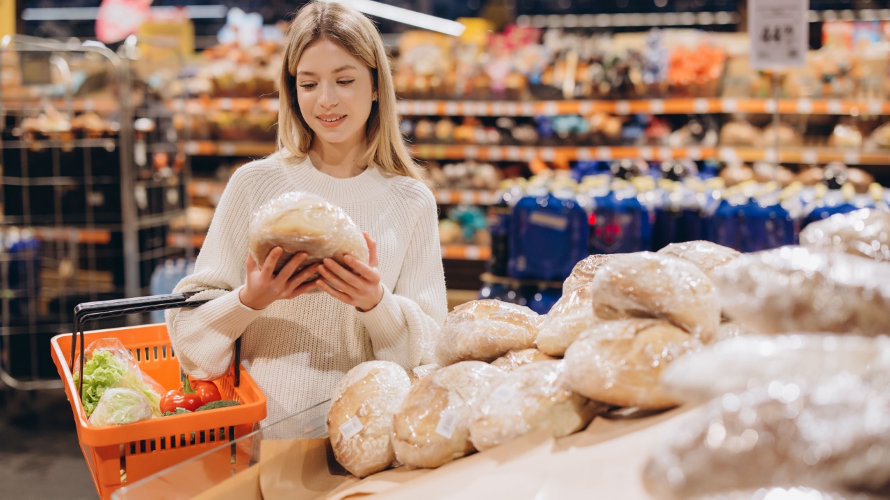 Customer selecting a loaf of bread from a supermarket display, holding a shopping basket with fresh produce