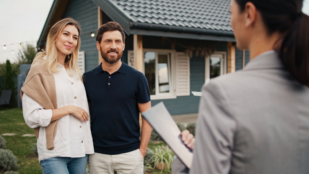 Caucasian husband and wife standing in front of beautiful small building. Talking with realtor. Man asking questions about price or possible additional information. Interested in buying.