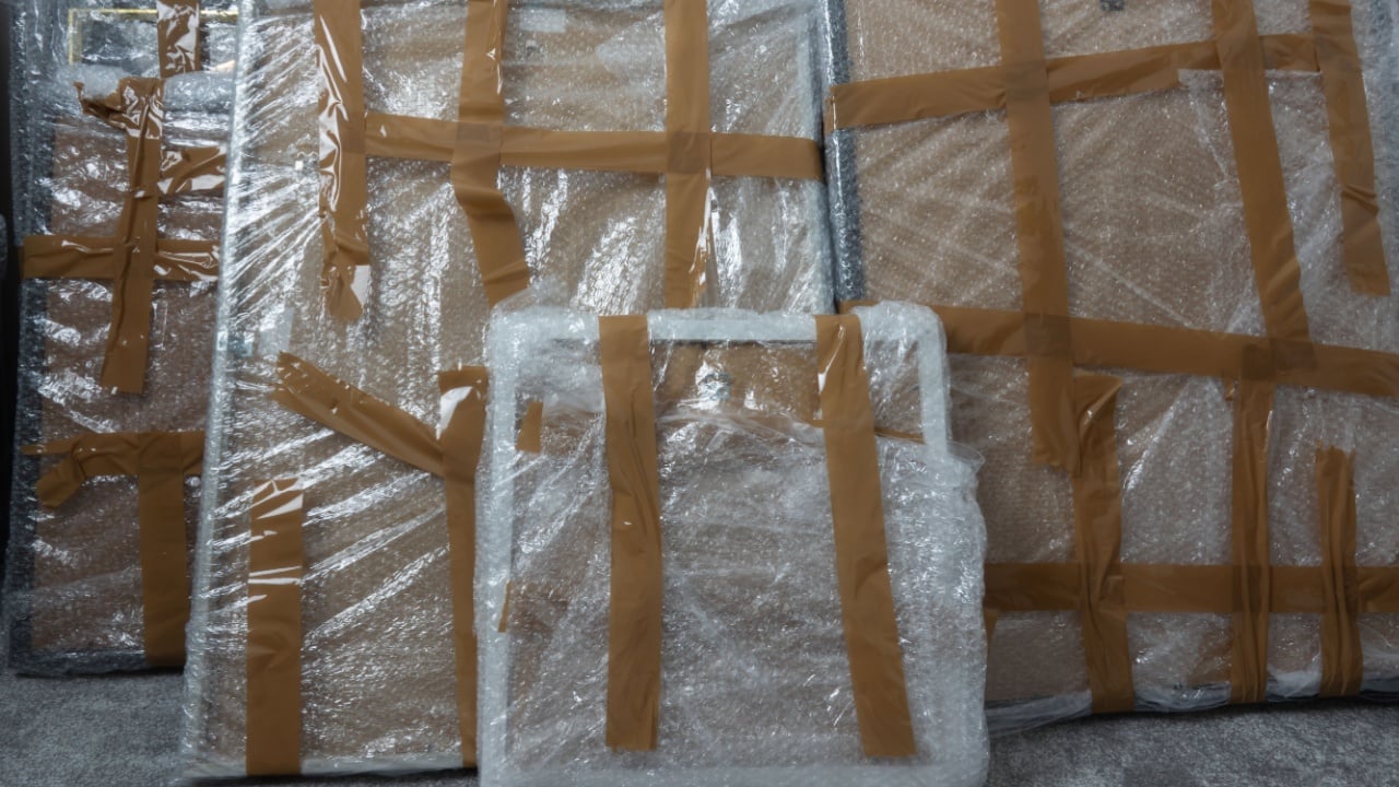 The image shows multiple paintings, all wrapped in bubble wrap and secured with brown tape. The paintings are of various sizes and are stacked on top of each other. 