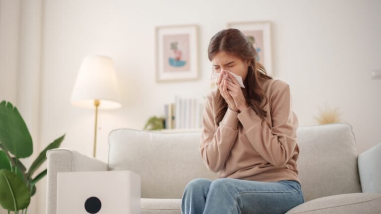 Young Asian woman sits on a couch, sneezing and holding a tissue to her nose while an air purifier operates nearby to improve air quality.