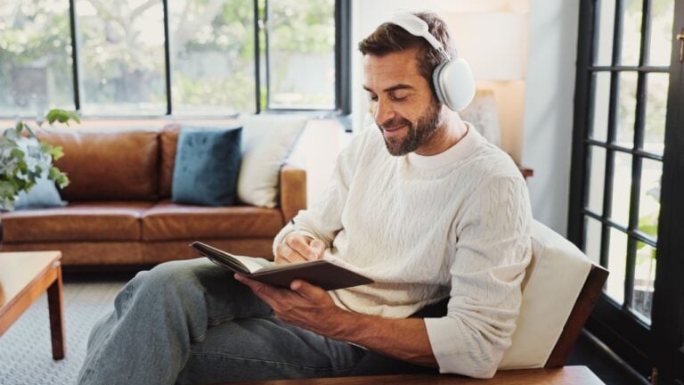 Home, man and smile with headphones for notebook, writing thoughts and personal reflection with music. Sofa, male person and happiness with audio track for journal, diary or to do list in living room