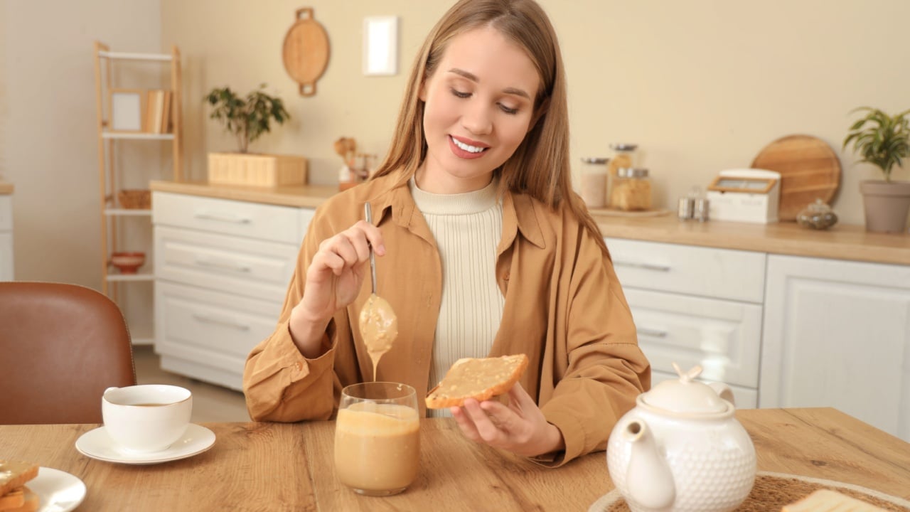 Young woman spreading peanut butter on toast with spoon near table in kitchen