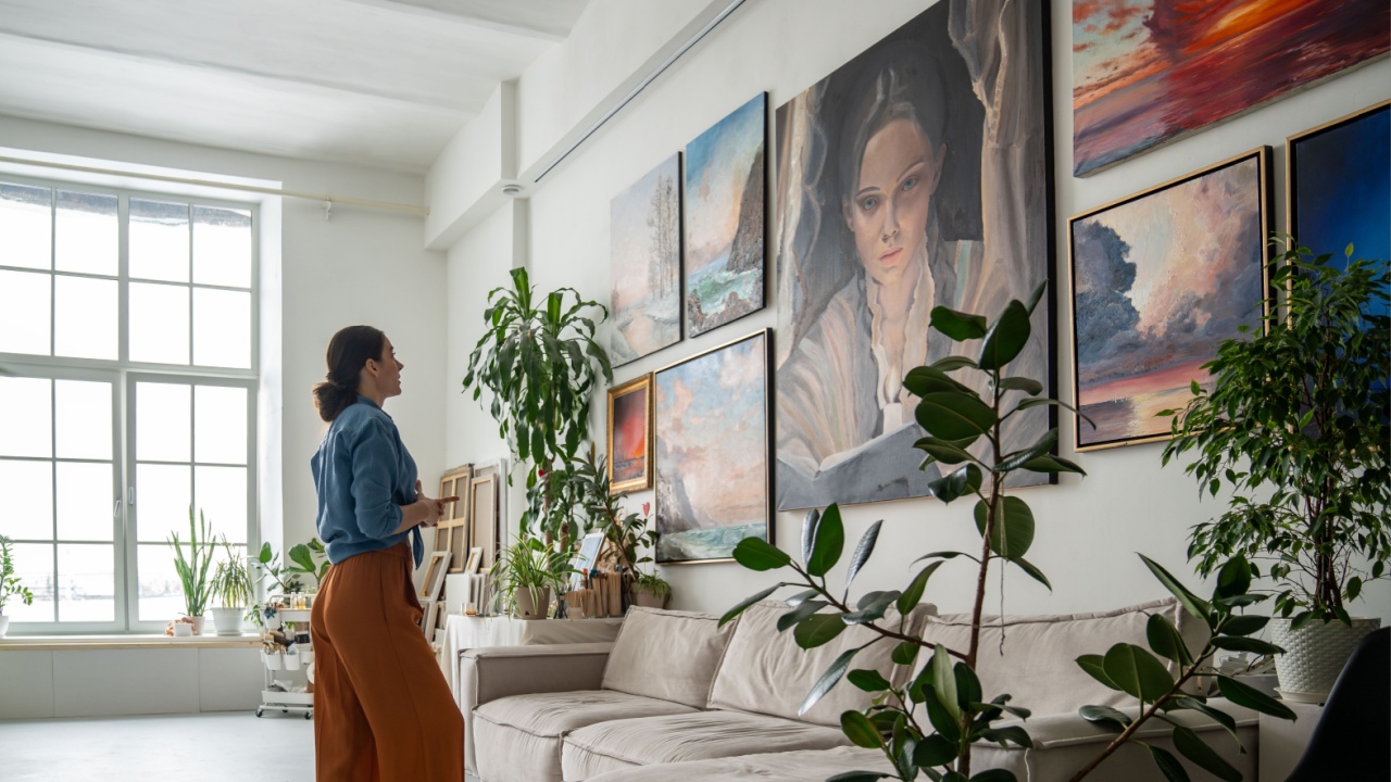 Female artist deeply reflects on portrait amidst other artworks on wall in studio. Creative workshop space with diverse collection of paintings, woman painter inspiration and new ideas for work