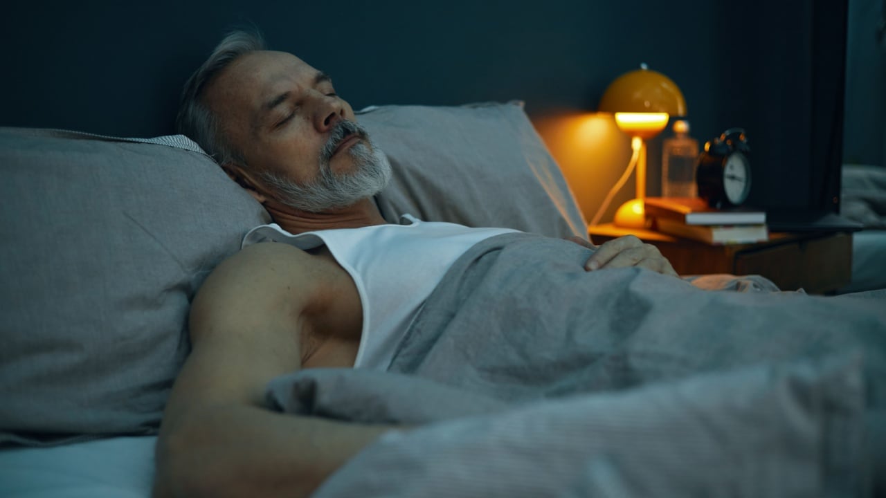 Elderly man peacefully sleeping in bed with warm lighting from bedside lamp illuminating scene. Clock and other bedroom objects visible on nightstand beside man