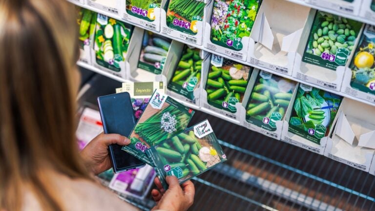 Woman holding vegetable seed packets and smartphone while choosing from shelf in gardening store. Sweden. Uppsala. 04.30.2025.