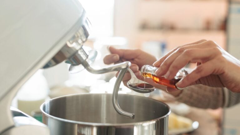 In a warm rustic kitchen, hands carefully add vanilla extract to a stand mixer, preparing to create fluffy Balkan Cozonac, a beloved Eastern European sweet bread filled with nuts and raisins