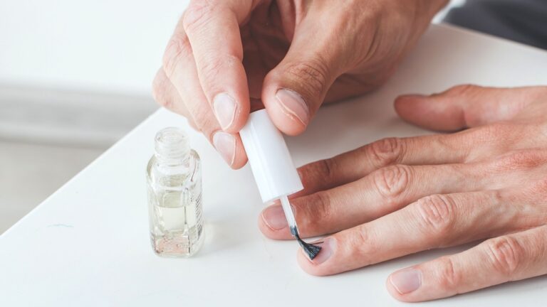 A man is applying clear nail polish to his fingernails at a white table. Only his hands, the brush, and the bottle are visible in the close-up shot.
