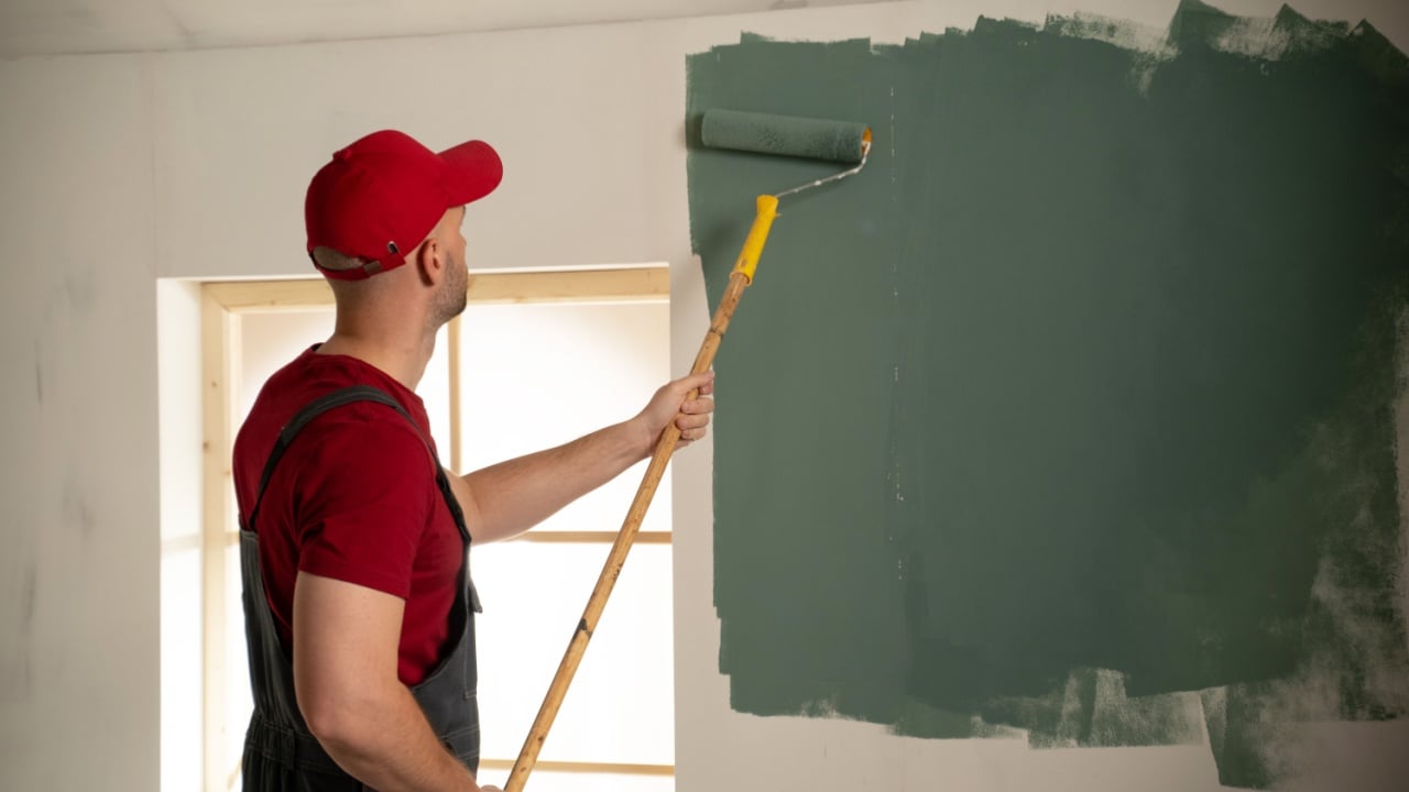 Professional painter wearing a red cap and overalls is skillfully applying green paint to a white wall using a long paint roller in a newly renovated house