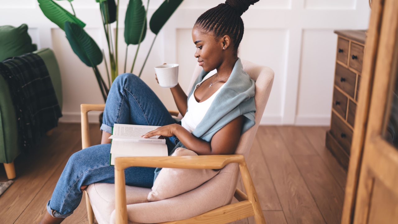 Side view of African American woman peacefully reading book and sipping coffee in cozy modern room with large plants and natural lighting, enjoying self-care moment in solitude