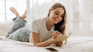 Young smiling caucasian woman writing in notebook while lying on bed in cozy living room