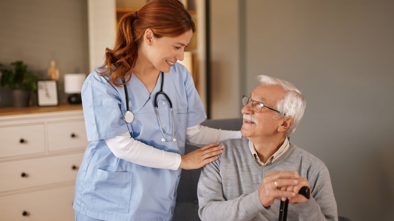 In a bright living room, a smiling, female home nurse in scrubs gently places her hand on the shoulder of an elderly gentleman. He sits holding a cane, looking up at her.