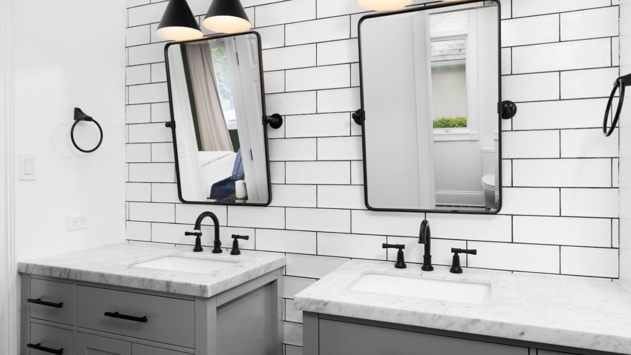 A bathroom detail with grey cabinets, white marble countertops, black faucets and light fixtures, and a white subway tile accent wall.