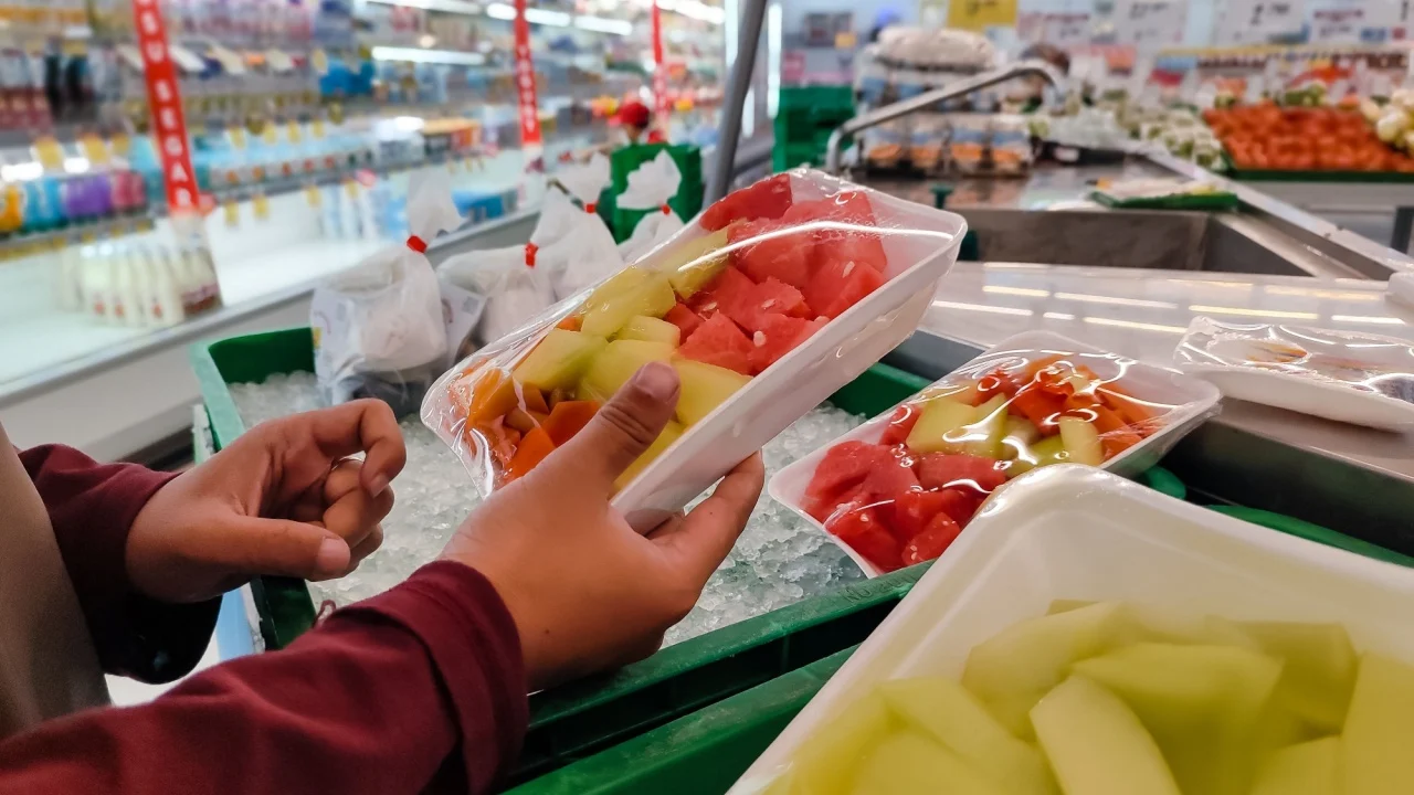 A customer selects a pack of freshly cut fruit from the chilled section of a supermarket. The fruits are neatly arranged in plastic trays, offering a convenient and healthy snack option.