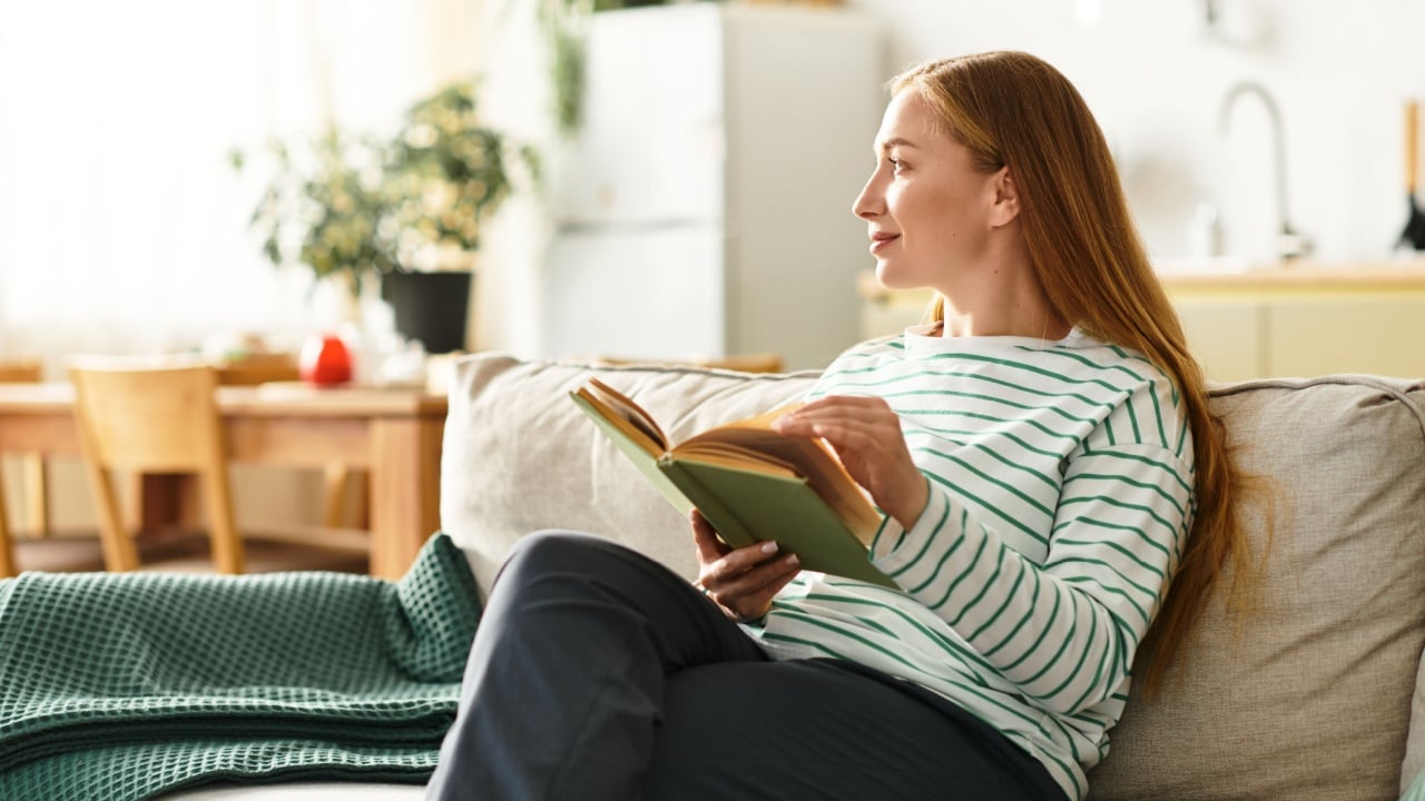 Young woman seated comfortably on a couch, deeply engaged in reading a book in her serene home.