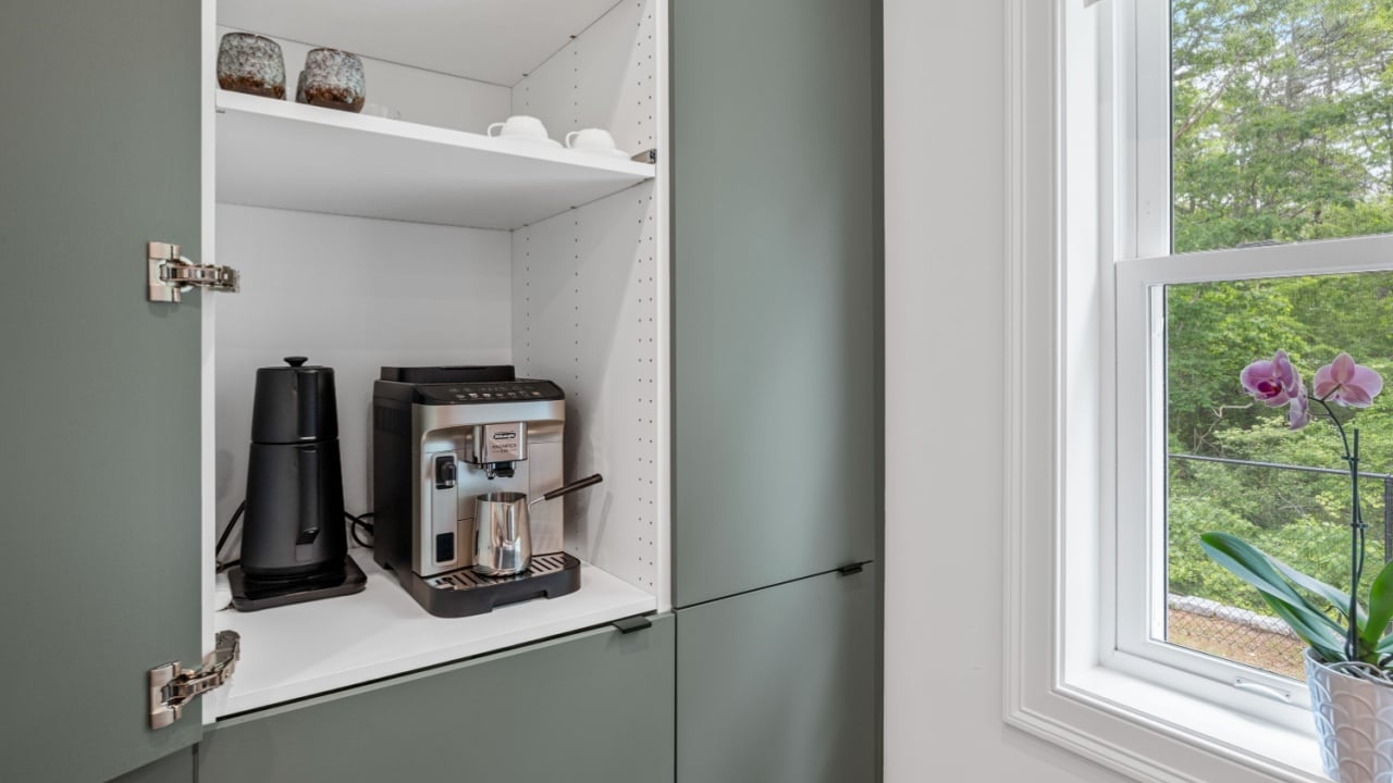 HARWICH, MASSACHUSETTS &ndash; JUNE 19, 2025: Built-in kitchen cabinet coffee station with espresso machine and milk frother beside a window with a flowering orchid plant.