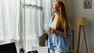 A young woman smiles contently while holding a warm drink, gazing out of the window at home.