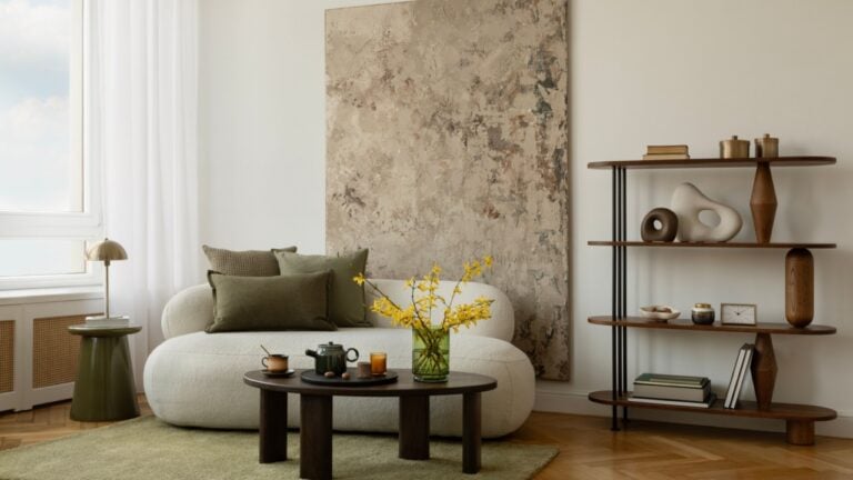 Minimalist living room with green-toned art, sculptural lamp, cozy white sofa, and a round coffee table with warm-toned decor and eucalyptus branches.