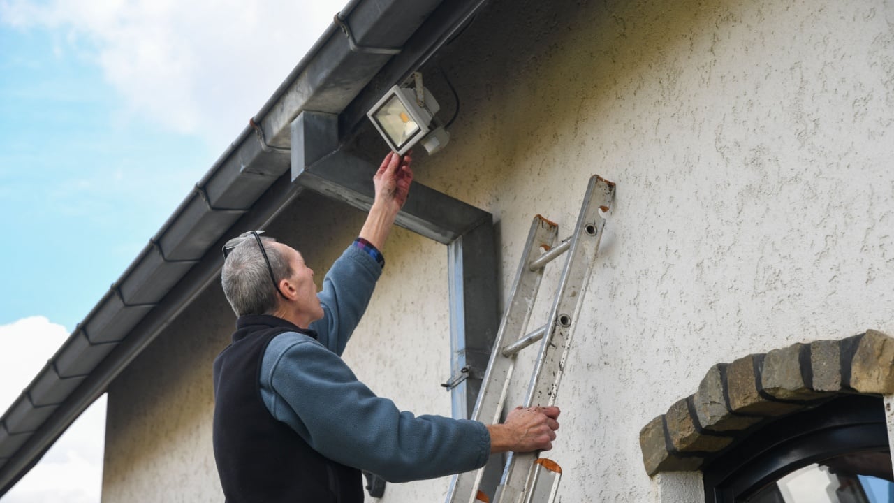 Electrician reaches for motion sensor LED floodlight while standing on ladder installing it on facade of house under gutter to improve outdoor lighting