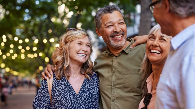 Group of happy senior friends sharing a moment outdoor while embrace. Older men and laughing women chatting together during a walk. Close up face of cheerful retirees enjoying in a lively city street.
