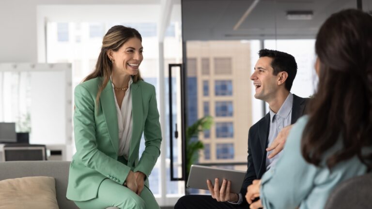 In stylish, modern office lounge area with city view, three young professionals engaged in relaxed, positive conversation, focus on smiling Hispanic woman laughing, enjoy interaction during briefing