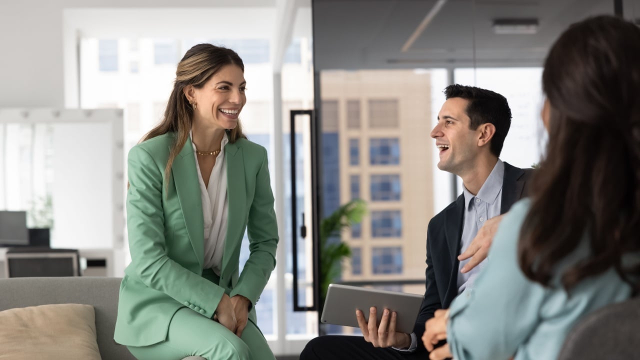 In stylish, modern office lounge area with city view, three young professionals engaged in relaxed, positive conversation, focus on smiling Hispanic woman laughing, enjoy interaction during briefing