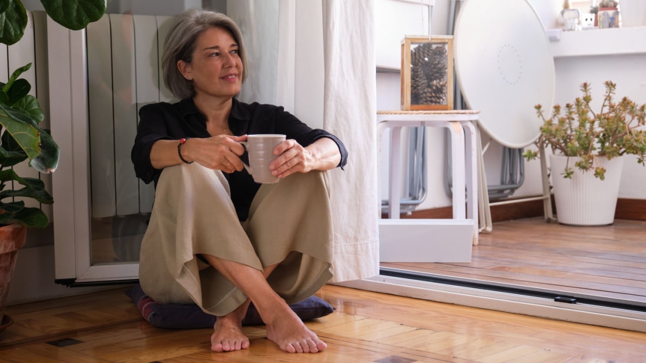 Serene mature woman enjoying a quiet moment with a cup of coffee, sitting on the floor by her balcony door, embracing the tranquility of her home